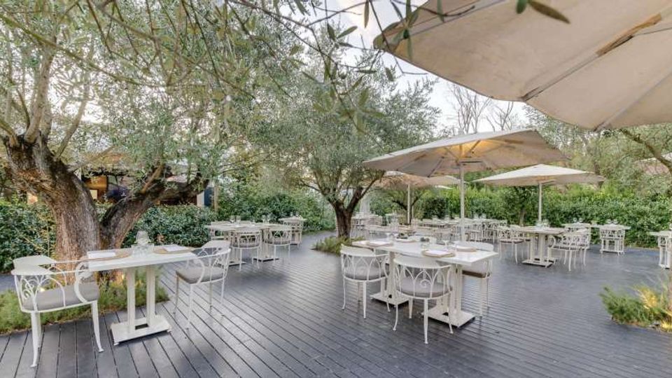 Terrazza ristorante all’aperto con pavimentazione in legno, tavoli e sedie bianchi e grandi ombrelloni. L’area è circondata da ulivi e siepi verdi, con vegetazione integrata tra i posti a sedere.