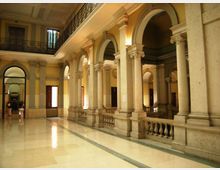 Ampio cortile interno di un edificio storico con pavimento in marmo lucido, porticato con alte colonne in pietra e grandi archi. Al livello superiore corre una balconata con ringhiera in ferro battuto, con finestre e porte lungo le pareti color ocra.