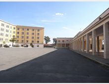 Ampio piazzale asfaltato tra edifici rettangolari a più piani con facciate color crema e molte finestre. Sul lato destro corre un lungo porticato con colonne in pietra che conduce verso un edificio basso sullo sfondo sotto cielo sereno.