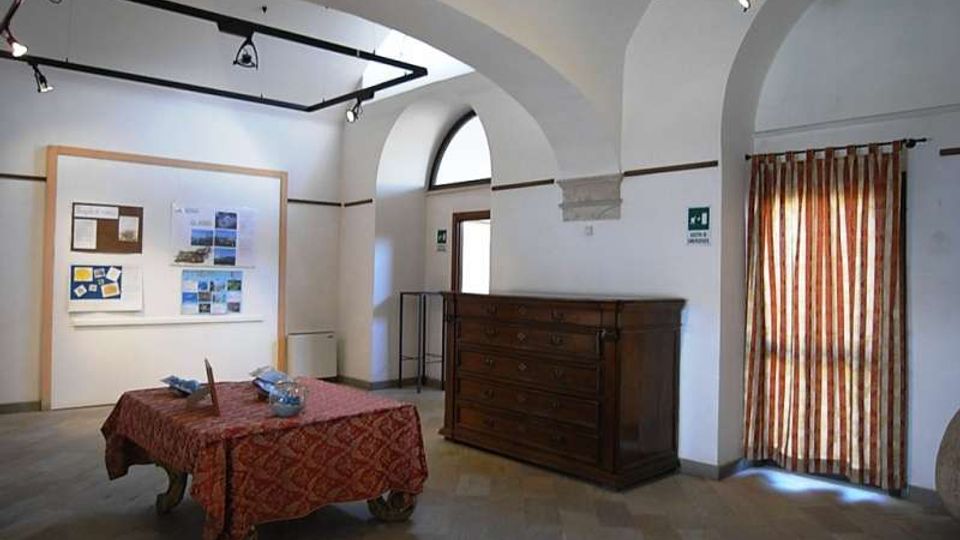 Interno di una piccola sala espositiva con soffitto a volte e archi bianchi, illuminazione a binari e pavimento in pietra. Al centro c’è un tavolo con tovaglia rossa, mentre lungo le pareti si vedono pannelli informativi, una grande cassettiera in legno e una finestra ad arco con tenda a righe.