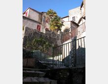 Scalinata in pietra in un borgo collinare, con ringhiere metalliche e muri in pietra ricoperti in parte da vegetazione. In alto si vedono case intonacate con persiane rosse e una palma sotto un cielo sereno.