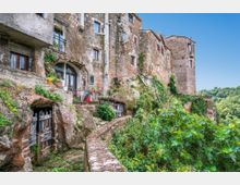 Borgo medievale con case in pietra addossate a una rupe, con facciate in muratura, finestre e archi, e porte ricavate nella roccia lungo un camminamento con muretto. Vegetazione rampicante e alberi circondano l’abitato, affacciato su una valle verde sotto un cielo sereno.