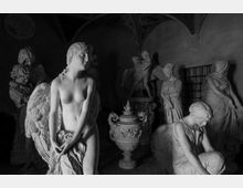 Interno di una galleria o deposito di sculture con statue in marmo disposte in un ambiente poco illuminato. In primo piano si vede una statua femminile con ali, mentre sullo sfondo compaiono altre figure classiche, un grande vaso decorato e archi con soffitto a volta.