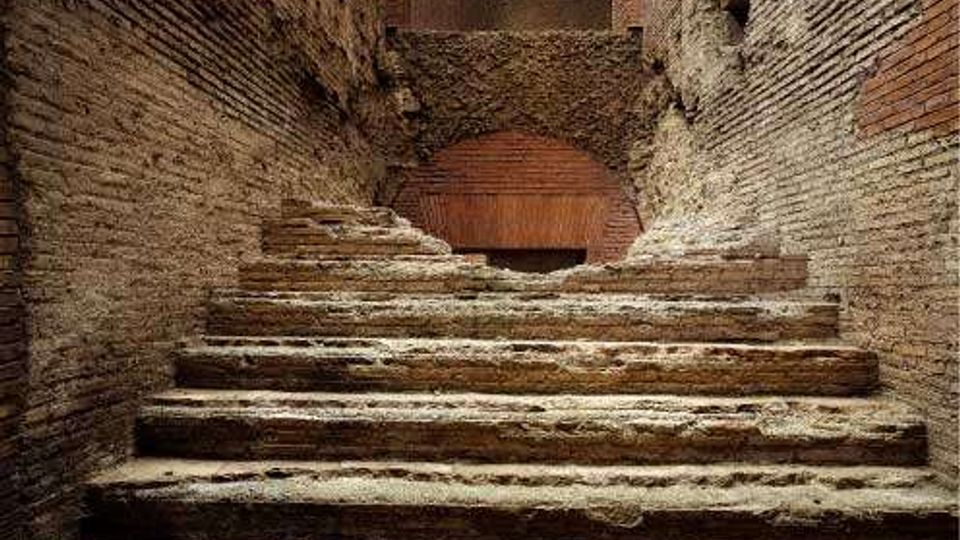 Scalinata in pietra consumata che sale tra strette pareti di mattoni e muratura antica. In cima si vede un arco in laterizio e un passaggio parzialmente chiuso da una muratura.