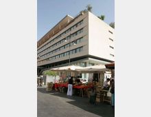 Edificio moderno a più piani con facciata chiara e lunghe finestre orizzontali, visto da un angolo su una strada urbana. In primo piano c’è un mercato all’aperto con bancarelle sotto grandi tende bianche, cassette di frutta e verdura e un lampione centrale.