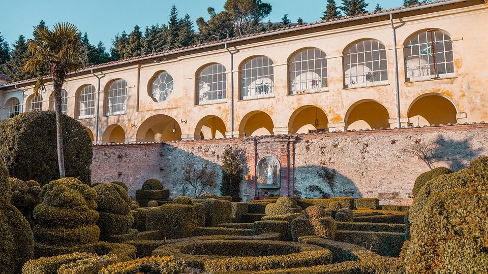Edificio storico intonacato con porticato ad archi e grandi finestre ad arco, affacciato su un muro in pietra. In primo piano un giardino formale con siepi potate in forme geometriche e un’alta palma, con alberi sullo sfondo.