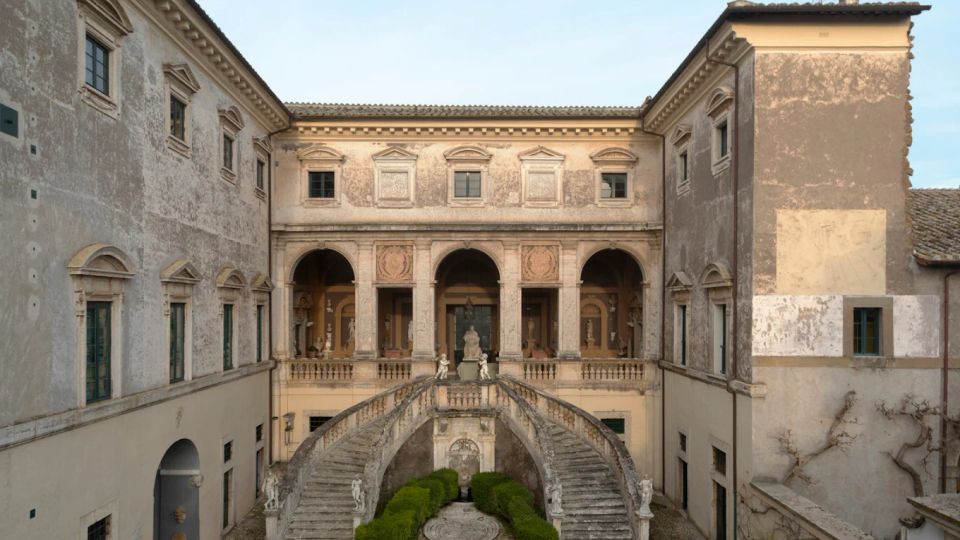 Cortile interno di un palazzo storico con facciate intonacate e finestre incorniciate, affacciato su una loggia ad archi. Al centro si trova una scenografica scalinata doppia in pietra con balaustre e statue, che scende verso un piccolo giardino geometrico con siepi e fontana.
