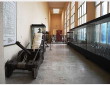 Corridoio museale lungo e stretto con alte finestre a griglia su un lato e grandi teche espositive in vetro. In primo piano si vede una grande macchina in legno e metallo tipo catapulta, con altri reperti e pannelli informativi lungo le pareti e una recinzione in fondo al passaggio.
