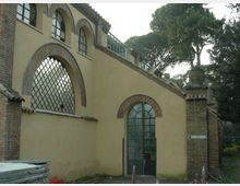 Facciata di edificio con intonaco chiaro e dettagli in mattoni, con grandi finestre ad arco e griglie metalliche. Accanto si vede un pilastro in mattoni con vaso decorativo e alberi alti sullo sfondo.