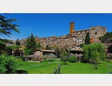 Veduta panoramica di un borgo medievale in pietra su una collina, con case addossate e una torre campanaria alta che domina il centro abitato. In primo piano si estende un prato verde con alberi e piccoli edifici rurali, sotto un cielo sereno.