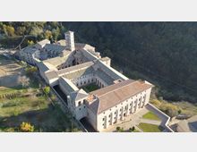 Veduta aerea di un grande complesso monastico in pietra con tetti in tegole, disposto attorno a un chiostro rettangolare e con un campanile quadrato centrale. L’edificio è circondato da aree verdi e da un bosco in collina, con cortili e vialetti lungo il perimetro.