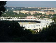 Veduta panoramica di un grande stadio ovale con struttura esterna bianca a traliccio, circondato da alberi e aree verdi. Sullo sfondo si estende la città con edifici e strade, osservata da un punto rialzato.