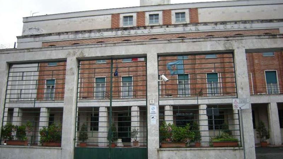 Facciata di un edificio pubblico in mattoni e pietra con portico a colonne e ampie griglie metalliche davanti alle finestre. In alto si vede una torretta con orologio e una fascia con scritta sulla facciata; davanti, cancellata e fioriere lungo l’ingresso.