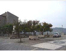 Piazzetta panoramica con pavimentazione in pietra e panchine in cemento sotto alcuni alberi, affacciata su un paesaggio collinare. Sul lato sinistro si vede un edificio storico in muratura e, sullo sfondo, una ringhiera in pietra con un lampione e un’auto parcheggiata.