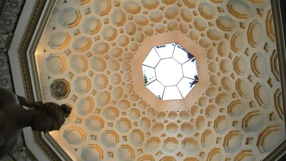 Vista verso l’alto di una cupola ottagonale decorata con motivi geometrici a cassettoni, con un lucernario poligonale centrale che lascia entrare la luce. Attorno si notano cornici e stucchi ornamentali lungo il perimetro della volta.