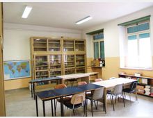 Aula scolastica con tavoli e sedie disposti in file, armadi vetrati pieni di libri lungo la parete e una carta geografica appesa. Grandi finestre con tapparelle verdi illuminano l’ambiente, con scaffali bassi e pile di libri vicino al davanzale.