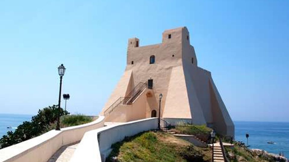 Fortezza costiera in muratura chiara su un promontorio, con alti muri inclinati e torre centrale. Un sentiero con muretto bianco e lampione conduce all’ingresso, con il mare sullo sfondo.