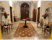 Interno di un ingresso in stile classico con soffitto a volta decorato, pavimento in marmo e inserto centrale in mattoni, e grande portone in legno ad arco sul fondo. Ai lati ci sono sedie e colonne con vasi di fiori, una piccola tavola rotonda al centro e un armadio in legno lungo la parete destra.