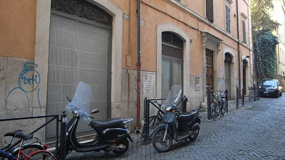Strada stretta in ciottoli affiancata da un edificio in intonaco color ocra con portoni e finestre ad arco, alcuni con inferriate. Lungo il marciapiede sono parcheggiati scooter e biciclette, con paletti e catene di protezione e un’auto sullo sfondo.
