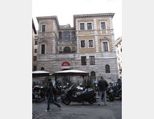 Facciata di un edificio storico in pietra e mattoni, con finestre rettangolari e cornicioni, identificato da un’insegna “Museo Barocco” sopra un ingresso ad arco. In primo piano si vede una strada urbana con molte moto parcheggiate e ombrelloni di un’area esterna.