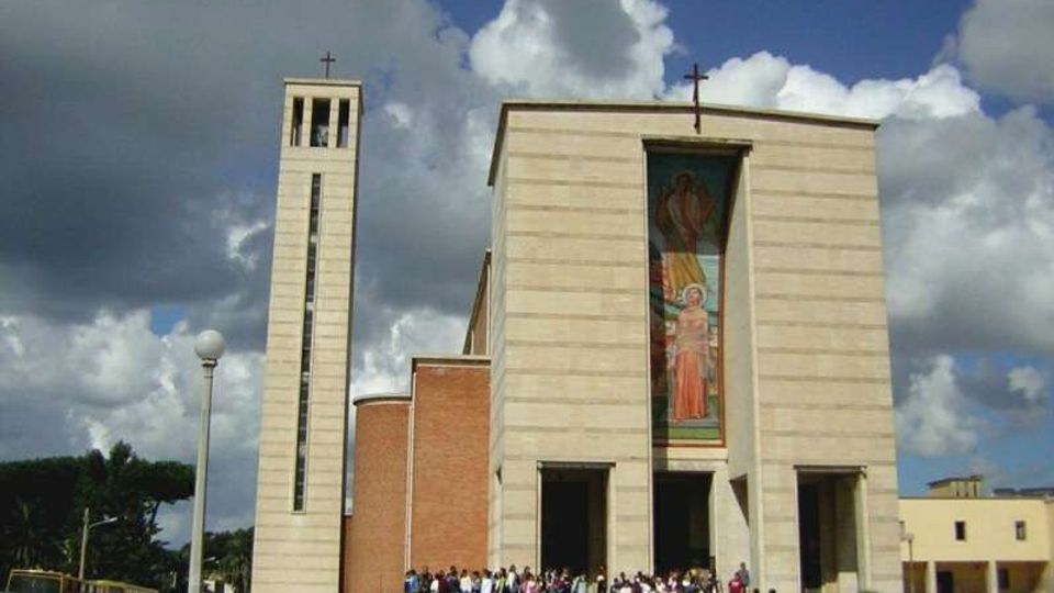 Facciata di una chiesa moderna in pietra chiara con grandi portali d’ingresso e un alto mosaico verticale al centro, affiancata da un campanile slanciato con croce in cima. Davanti si apre una piazza con auto parcheggiate e lampioni, sotto un cielo nuvoloso.