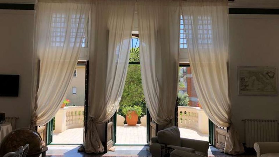 Interno di un salone luminoso con tre alte porte-finestre ad arco, coperte da tende bianche, che si aprono su una terrazza con balaustra in pietra e un grande vaso. Pavimento piastrellato decorato e arredi da soggiorno con divani; all’esterno si vede vegetazione e alcuni edifici vicini.