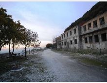 Strada sterrata lungo una scogliera, con alberi e panchine sul lato sinistro e un lungo edificio in pietra abbandonato con finestre vuote sul lato destro. Sullo sfondo si intravede il mare e l’orizzonte al tramonto.