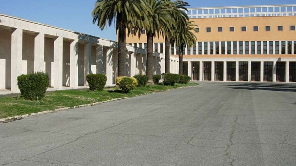 Ampio viale asfaltato che conduce a un complesso architettonico moderno con facciate chiare, un porticato con pilastri sul lato sinistro e un edificio principale con grandi finestre e ingressi vetrati. Palme alte e siepi basse fiancheggiano l’area verde lungo il percorso.