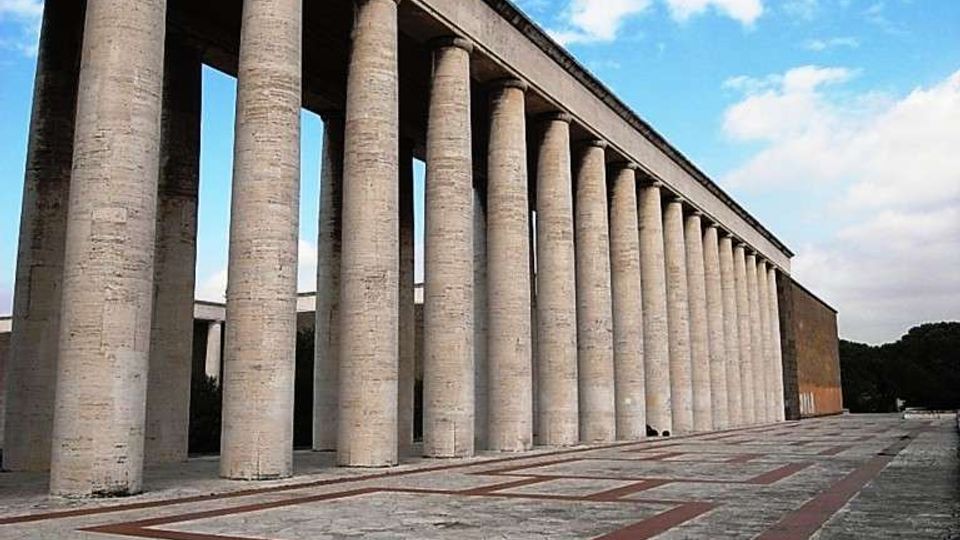 Lungo colonnato monumentale in pietra con alte colonne cilindriche che sorreggono un’ampia trabeazione, affacciato su una grande piazza pavimentata con motivi geometrici. Sullo sfondo si vede un cielo azzurro con nuvole e una fascia di alberi ai margini.