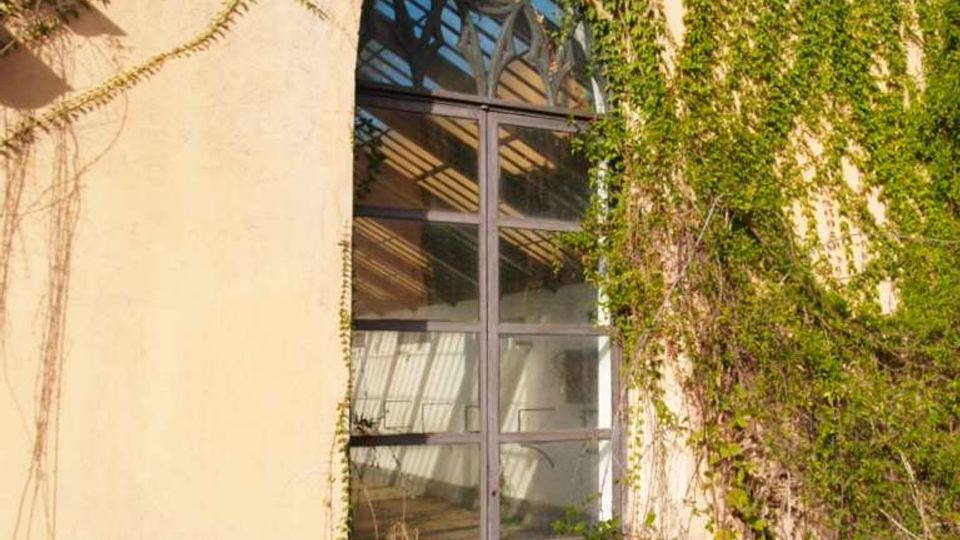 Facciata di edificio chiaro con grande finestra ad arco a vetri, incorniciata da una struttura metallica. La parete e il terreno davanti sono ricoperti da edera e vegetazione rampicante che scende dall’alto.