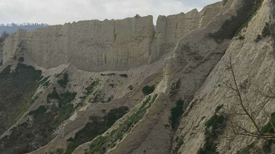 Ripida parete di falesia rocciosa e friabile, con creste erose e canaloni verticali, punteggiata da piccoli cespugli. Sullo sfondo si vedono altre formazioni collinari sotto un cielo nuvoloso.