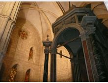 Interno di una chiesa in pietra con alte volte a crociera, archi gotici e finestre strette con vetrate colorate. In primo piano si vede un grande baldacchino scuro con colonne e arco decorato vicino all’altare.