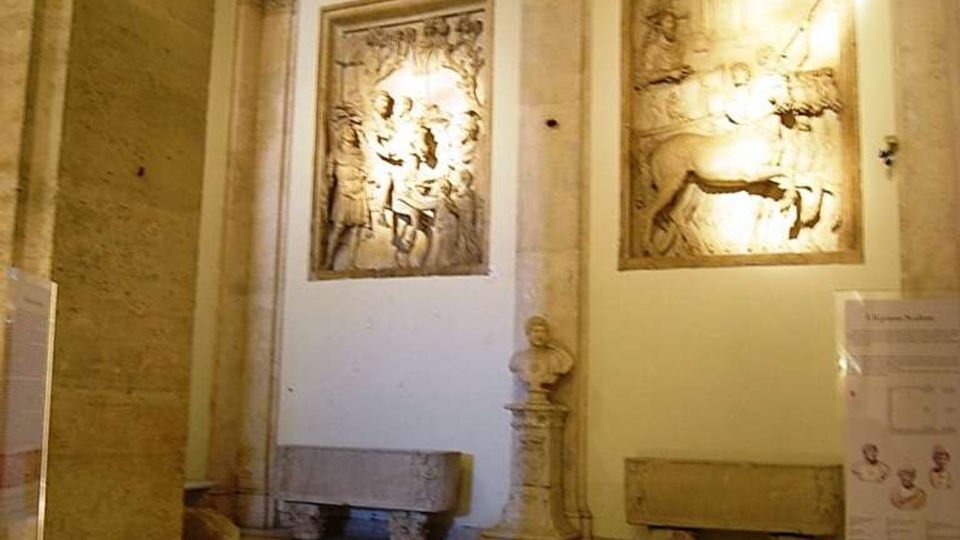 Interno di una galleria o museo in pietra, con colonne e cornici classiche e pavimento a riquadri. Sulla parete sono esposti due grandi bassorilievi illuminati, con panche in pietra e un busto su piedistallo tra le colonne.