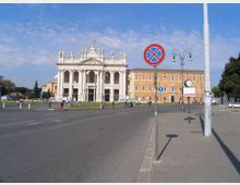 Ampia piazza urbana con strada asfaltata e marciapiede in primo piano, segnaletica di divieto di sosta e lampioni. Sullo sfondo si vede una grande basilica in stile neoclassico con facciata bianca a colonne e statue, affiancata da edifici storici color ocra.