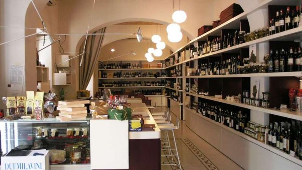 Interno di un’enoteca con lunghi scaffali a parete pieni di bottiglie e prodotti, bancone centrale e vetrina refrigerata in primo piano. Soffitto con travi in legno e archi, illuminato da lampade a sospensione sferiche, con corridoio decorato che conduce verso il fondo del negozio.