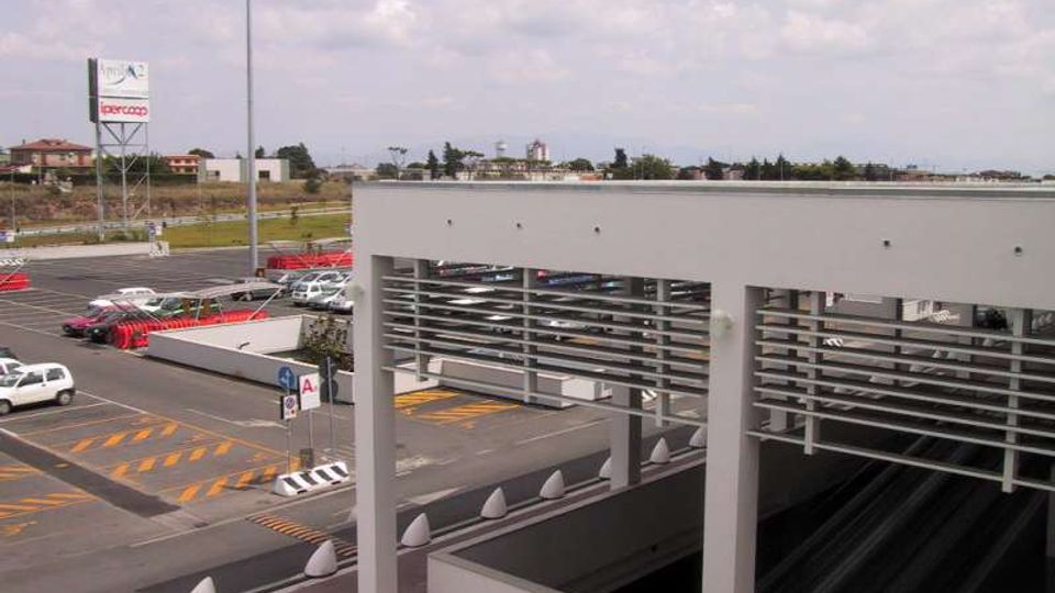 Area esterna di un grande centro commerciale con ampio parcheggio e corsie di accesso segnalate, con auto e lampioni alti. In primo piano una struttura moderna bianca con pilastri e pannelli orizzontali copre una rampa che scende verso un livello sotterraneo.