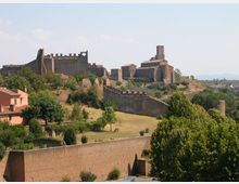 Veduta panoramica di un borgo collinare con un castello medievale in pietra e una grande chiesa fortificata con torre, circondati da mura merlate. In primo piano si vedono campi e alberi, con una lunga cinta muraria che attraversa il paesaggio.