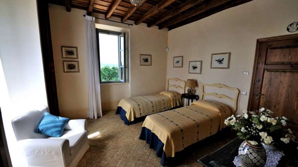 Camera da letto con due letti singoli affiancati, soffitto con travi in legno a vista e pavimento in piastrelle. Una finestra con tenda chiara illumina la stanza, arredata con poltrona bianca, comodino con lampada, quadri alle pareti e porta in legno.
