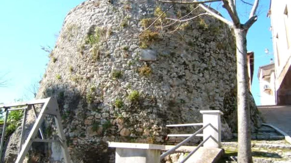 Grande struttura circolare in pietra grezza, simile a una torre o bastione fortificato, vista dal basso. In primo piano ci sono scale e muretti in cemento con ringhiere, un albero spoglio e un’area in pendenza accanto a edifici.