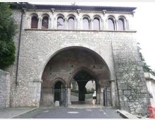 Facciata in pietra di una porta monumentale con grande arco centrale che attraversa un passaggio stradale, affiancato da due ingressi ad arco con cancelli in ferro. Sopra l’arco si trova una fila di finestre ad arco in stile romanico sotto un tetto con sporto, con alberi ai lati.