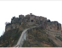 Borgo storico in pietra arroccato sulla sommità di una rupe di tufo stratificato, con edifici compatti e una chiesa con piccolo campanile. Una lunga strada sopraelevata con parapetti sale dal basso fino all’ingresso del paese lungo il crinale della scogliera.