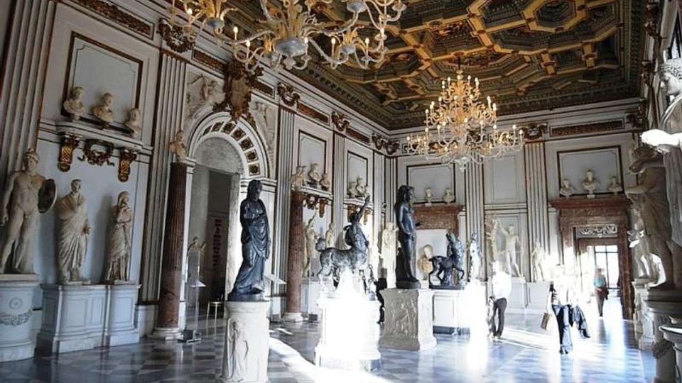 Ampia sala museale in stile neoclassico con pavimento a scacchi, pareti ornate da nicchie con statue e busti, e grandi lampadari di cristallo sospesi sotto un soffitto riccamente decorato. Al centro della stanza sono esposte sculture su piedistalli, con luce naturale che entra da ampie porte e finestre.