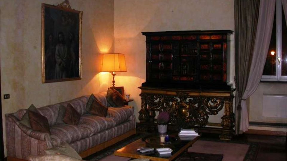 Salotto in stile classico con divani imbottiti, tavolino basso e tappeti su pavimento in legno, illuminato da una lampada da terra. Sulla parete si vede un grande dipinto e, di fronte, un mobile antico intagliato accanto a una finestra con tende lunghe.