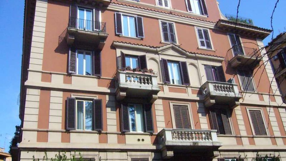 Facciata di un palazzo residenziale di cinque piani con intonaco color terracotta e cornici chiare, finestre con persiane scure e più balconi sporgenti con ringhiere in ferro e balaustre. L’edificio è ripreso dal basso contro un cielo azzurro, con alcuni rami e vegetazione visibili ai margini dell’inquadratura.