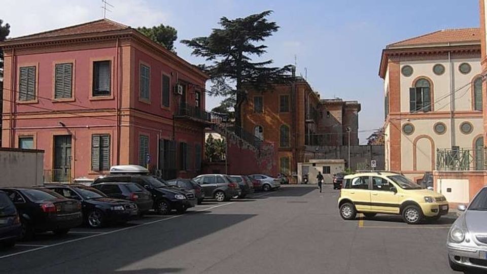 Ampio parcheggio asfaltato tra edifici bassi in stile tradizionale con facciate colorate e finestre con persiane. Sullo sfondo si vedono altri fabbricati e un grande albero, con auto parcheggiate lungo i lati sotto un cielo sereno.