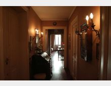 Corridoio interno di un appartamento d’epoca con pareti color ocra, pavimento in legno e applique a parete in stile classico. Ai lati si vedono porte e piccoli arredi con specchi e quadri, mentre in fondo una finestra illumina una stanza adiacente.