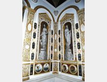 Interno di una sala decorata con stucchi dorati e pannelli ornamentali, con due grandi nicchie ad arco contenenti figure classiche in rilievo. Le pareti sono arricchite da cornici e medaglioni dipinti con motivi naturalistici lungo la parte inferiore.