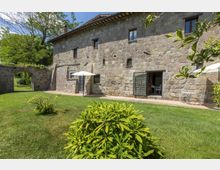Cortile erboso con arbusti davanti a un edificio rurale in pietra a due piani, con piccole finestre e porte vetrate che si aprono su un patio. Sulla terrazza ci sono tavoli e sedie con ombrelloni bianchi, e a sinistra un arco in pietra conduce a un’area verde.