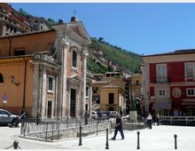 Piazza di un centro storico con una piccola chiesa in pietra con facciata rettangolare, portale centrale e croce sul tetto, circondata da ringhiera metallica. Sullo sfondo si vedono edifici colorati e case sul pendio di una collina verde sotto un cielo sereno.