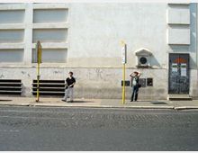 Marciapiede lungo una strada asfaltata davanti a un grande edificio con facciata chiara, riquadri rettangolari in rilievo e segni di graffiti nella parte bassa. Sono visibili due paline gialle di fermata, una griglia di aerazione e una porta scura con adesivi e scritte.